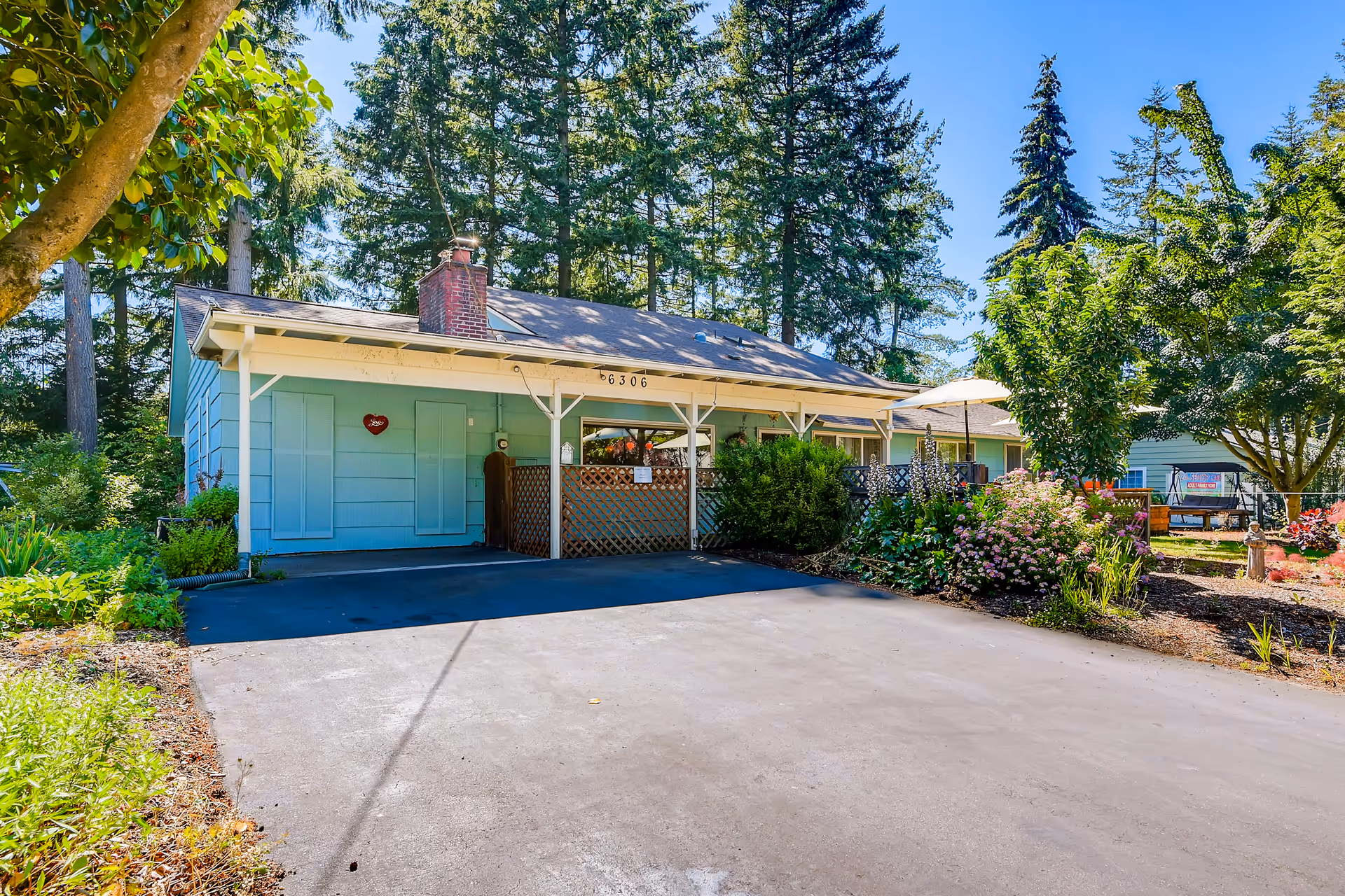 A single-story light blue house with a covered porch and a brick chimney, surrounded by lush greenery and trees under a clear blue sky. There is a paved driveway in front and a garden with various plants and flowers on the right side.