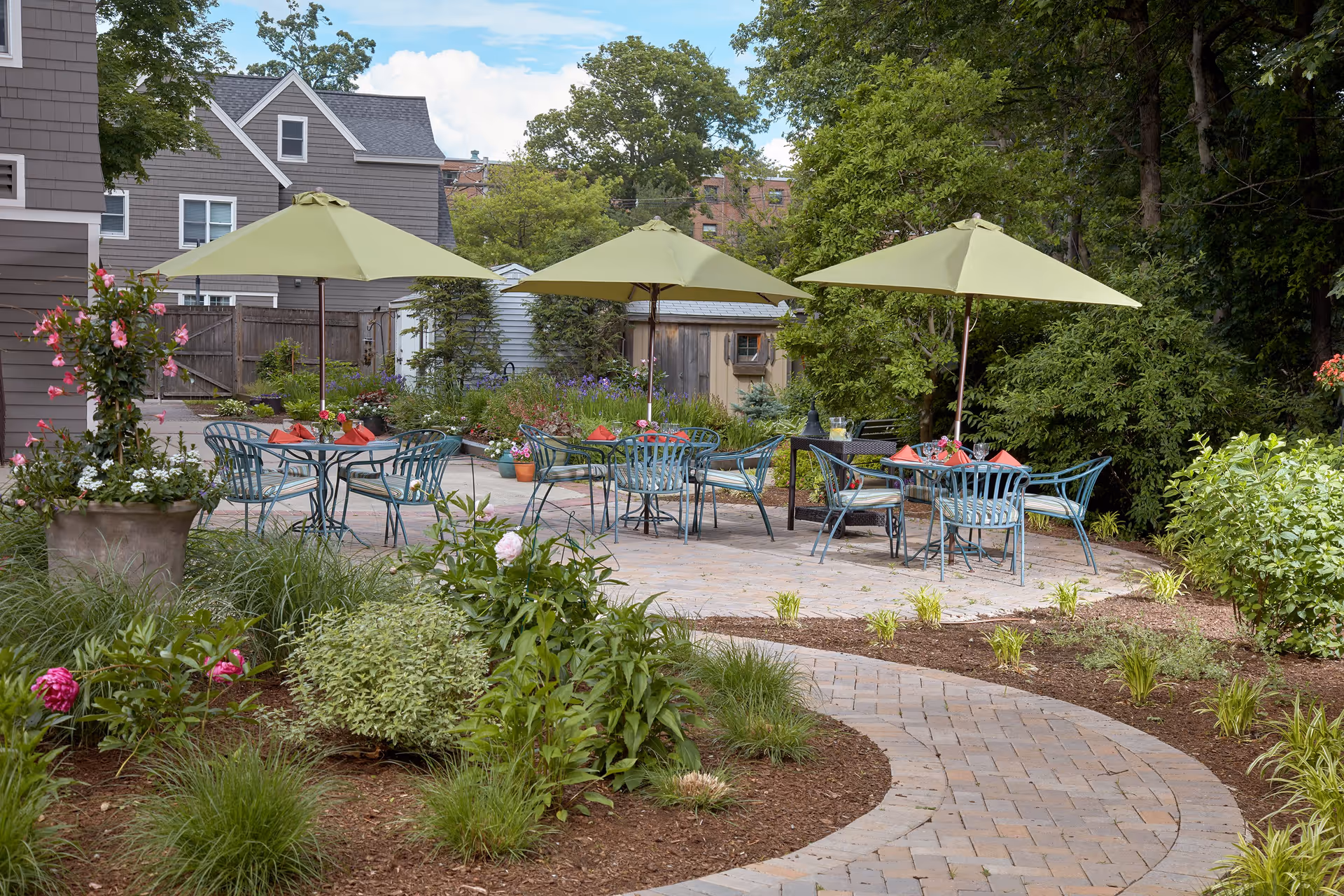 Outdoor patio area with three green umbrellas shading metal tables and chairs, surrounded by lush greenery, flowering plants, and a curved brick pathway leading to the seating area. Residential buildings and a wooden fence are visible in the background.