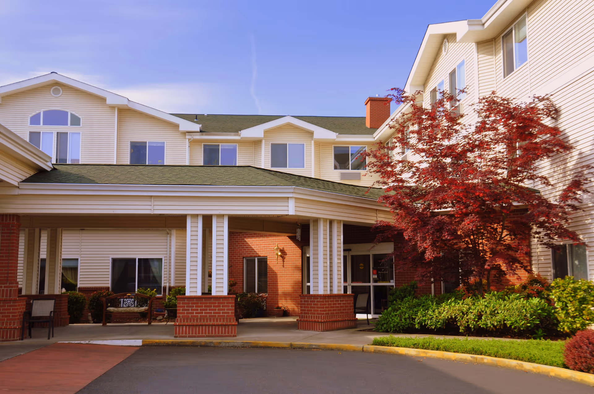 Exterior view of a senior living facility building with beige siding and green roof. The entrance has a covered drop-off area supported by white columns with red brick bases. There is a red-leafed tree and green shrubs near the entrance, and a clear blue sky above.