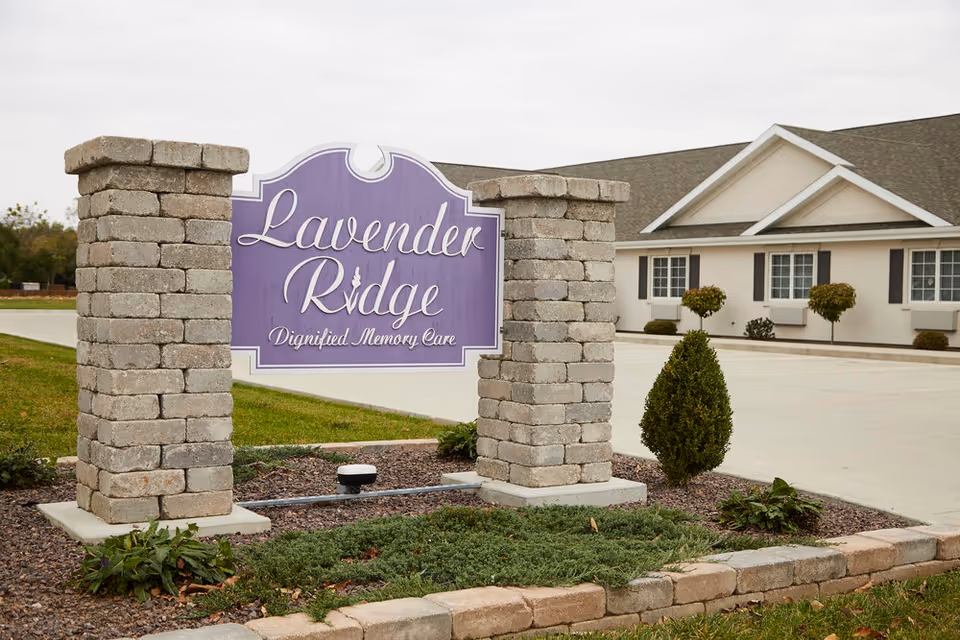 Stone sign for Lavender Ridge Dignified Memory Care mounted between two stone pillars with a building and driveway in the background under an overcast sky.