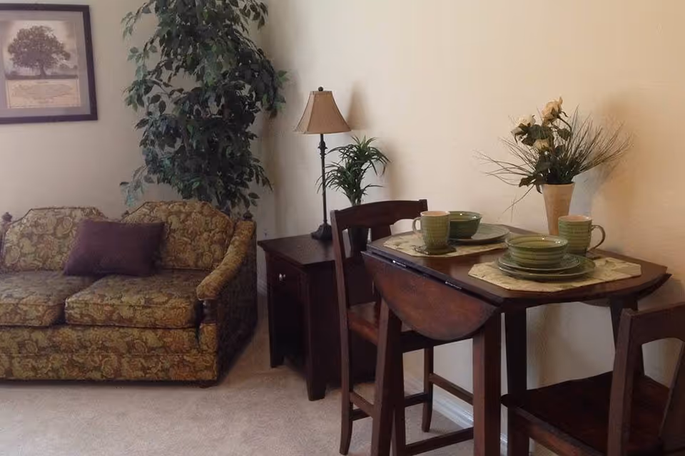 A cozy living area featuring a patterned sofa with a purple cushion, a small wooden side table with a lamp and a potted plant, and a wooden dining table set for two with green dishes and a vase of flowers against a plain light-colored wall.