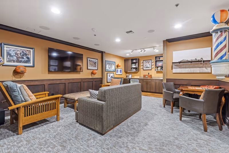 A cozy living room area with a gray couch, wooden armchair, and ottoman facing a wall-mounted flat screen TV. The walls are decorated with framed sports memorabilia and basketballs. There is a small table with two chairs on the right side, and built-in wooden cabinets with shelves displaying various items. The room has carpeted flooring and warm lighting.