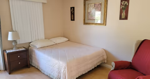 A bedroom with a neatly made bed covered with a light pink quilt. There is a wooden nightstand with a lamp and a book on it next to the bed. The walls are beige with three framed floral artworks hanging above the bed. A red upholstered armchair is positioned to the right of the bed.