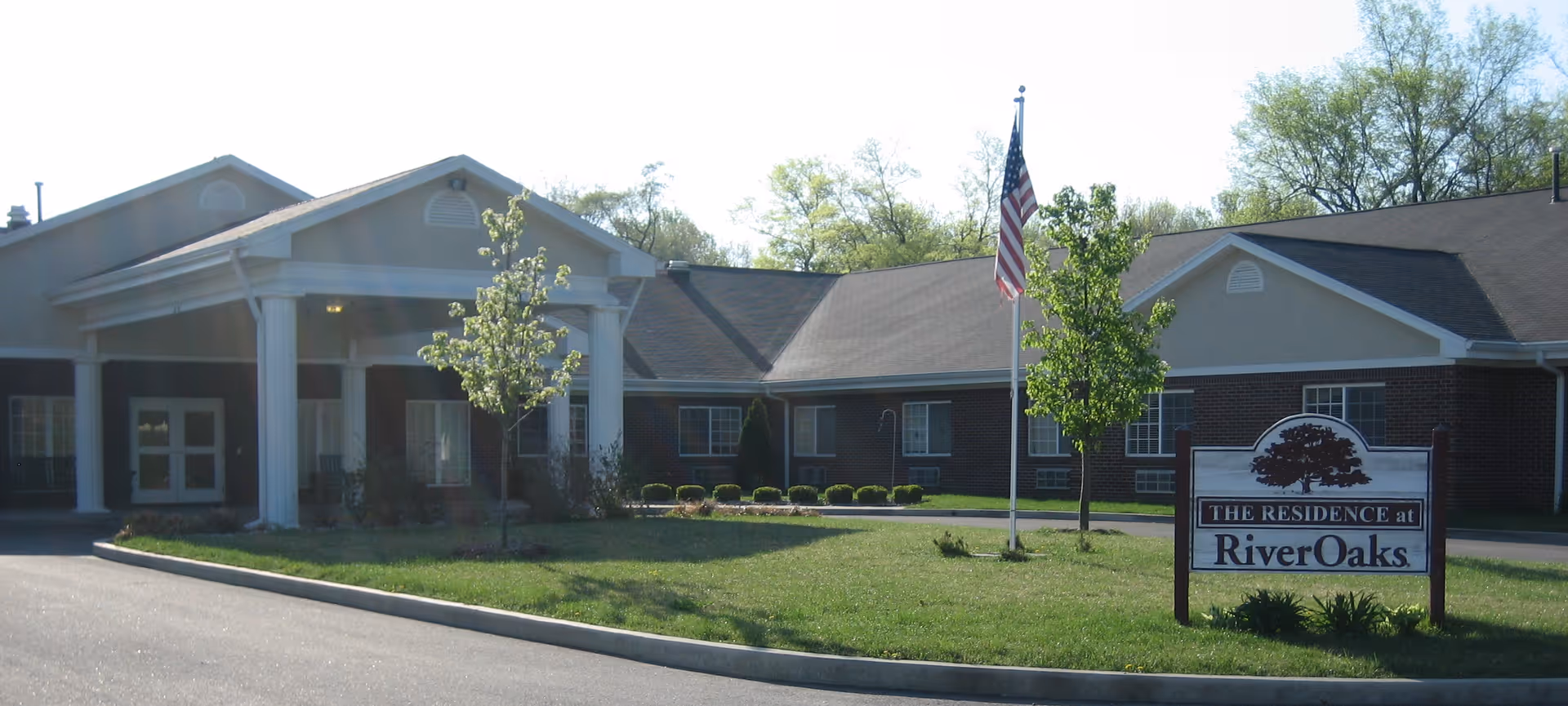 Exterior view of RiverOaks Health Campus showing a single-story brick building with white columns at the entrance, a flagpole with an American flag, and a sign that reads 'THE RESIDENCE at RiverOaks' on a grassy area.