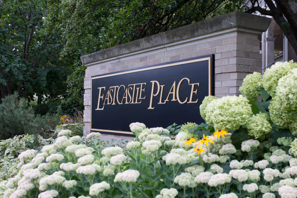 A brick sign with a black background and gold lettering that reads 'Eastcastle Place,' surrounded by green foliage and white and yellow flowers.