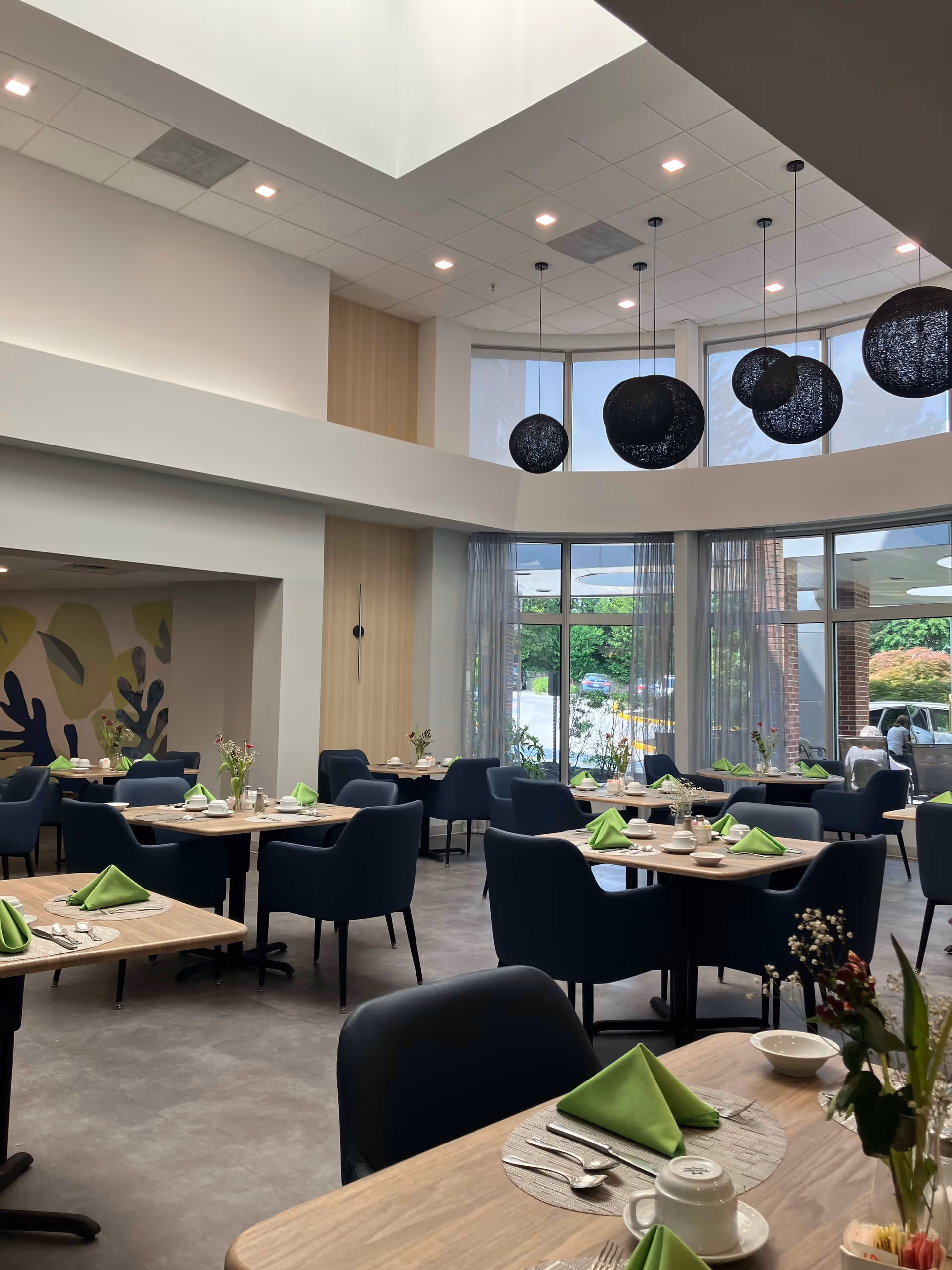 A bright and modern dining room with multiple wooden tables set with green folded napkins, white cups, and silverware. The room features large windows with sheer curtains, dark blue upholstered chairs, and several black spherical pendant lights hanging from the ceiling. There is a colorful abstract mural on one wall and a view of the outdoor parking area through the windows.