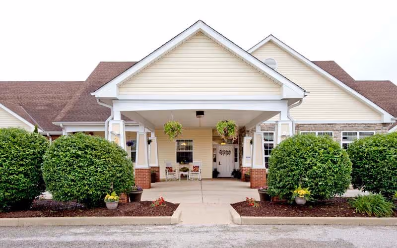 Front exterior view of a single-story building with a covered entrance supported by white columns. The building has beige siding, a brown shingled roof, and neatly trimmed round green bushes along the walkway leading to the entrance. Two hanging flower baskets are suspended from the covered entrance ceiling, and two chairs with cushions are placed near the door.