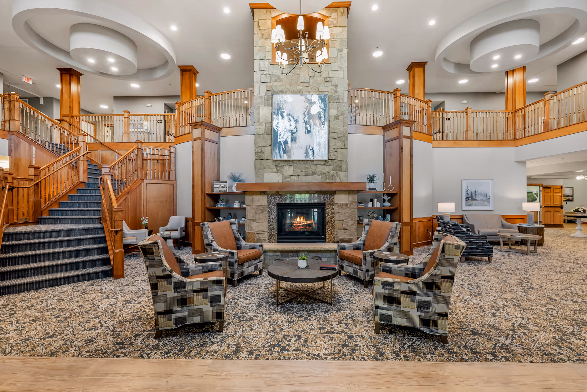 Spacious and elegant living room area in Cherrywood Pointe Assisted living facility featuring a large stone fireplace with a modern painting above it, surrounded by comfortable patterned and leather chairs arranged around a round coffee table. The room has wooden railings on the upper level, carpeted floors, and a grand staircase on the left side. Warm lighting and tasteful decor create a welcoming atmosphere.