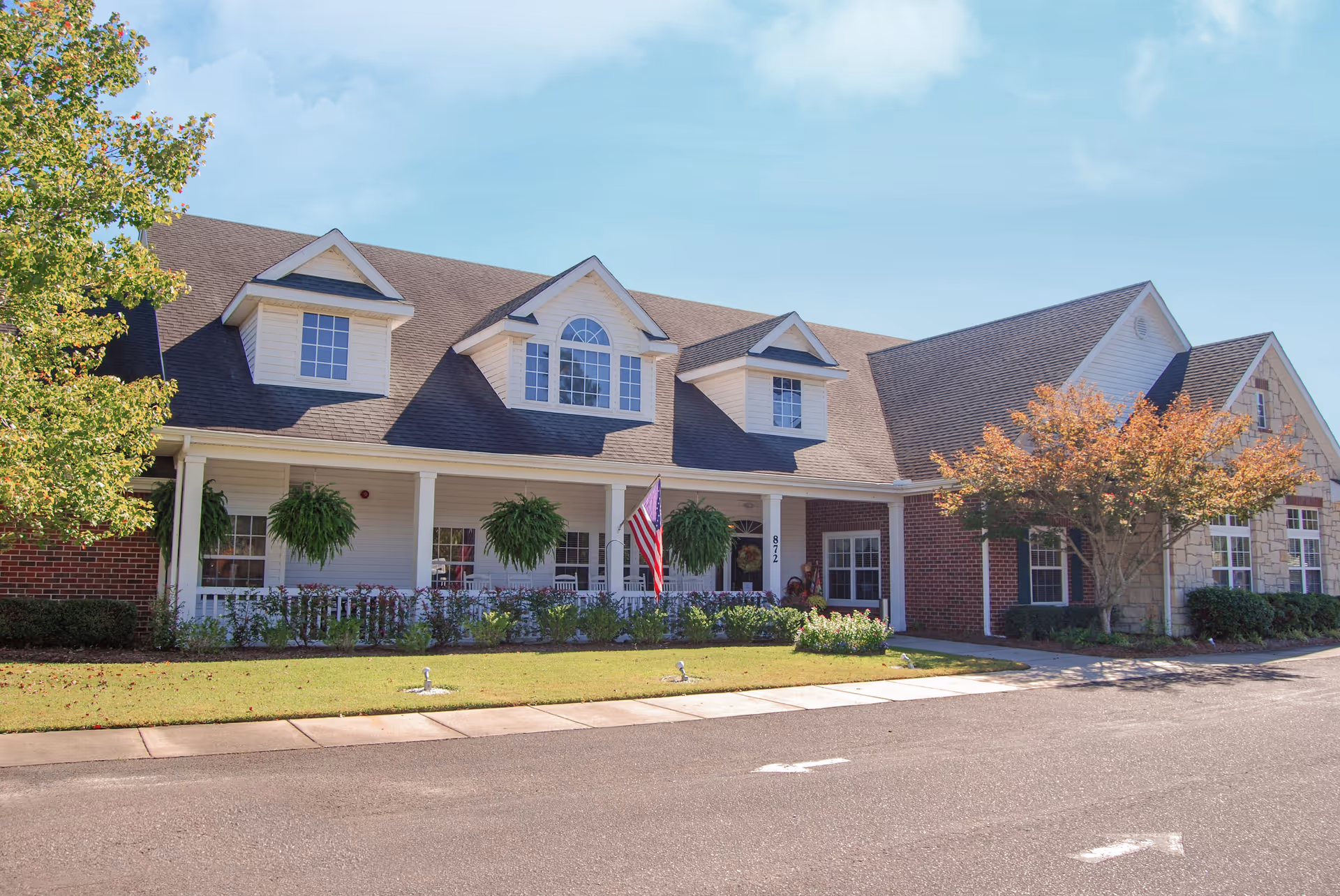 Exterior view of a senior living facility building with a porch featuring hanging plants and rocking chairs. The building has a combination of brick and stone walls, multiple windows, and a sloped roof with dormer windows. There is an American flag displayed on the porch and a well-maintained lawn with shrubs and trees in front.