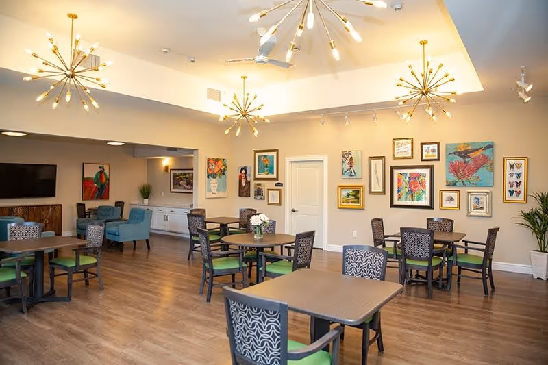 A bright and spacious common area in a senior living facility with multiple tables and chairs arranged for seating. The room features modern light fixtures hanging from the ceiling, a wall adorned with colorful framed artwork, and a flat-screen TV mounted on the wall. The floor is wooden, and there are plants placed around the room for decoration.