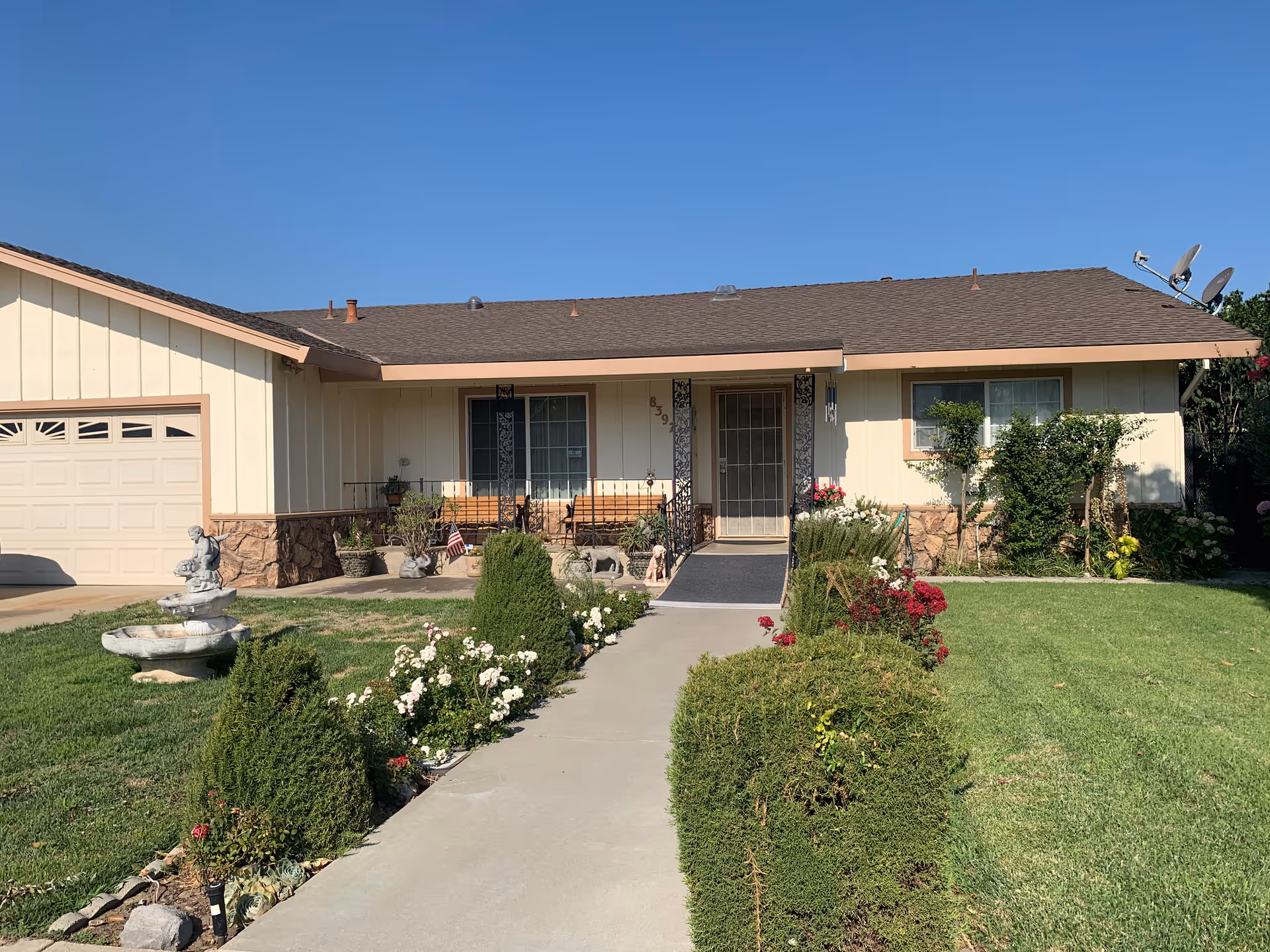 Single-story house with a beige exterior and brown roof, featuring a front porch with a bench and decorative plants. A concrete walkway leads from the sidewalk to the front door, bordered by neatly trimmed bushes and flowering plants. The lawn is green and well-maintained under a clear blue sky.