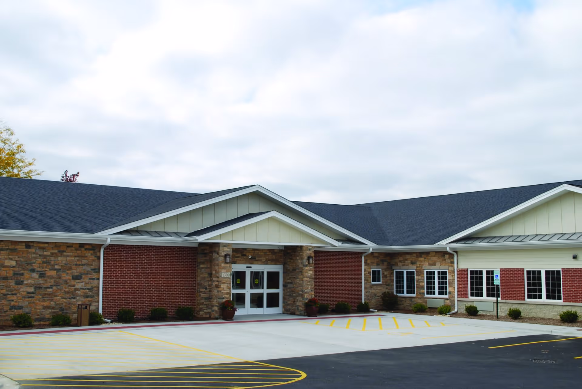 Exterior view of a single-story senior living facility building with a combination of brick and stone facade, white trim, and a dark shingled roof. The entrance has double glass doors under a small gabled roof. There is a parking area with marked spaces and a handicapped parking sign near the entrance.