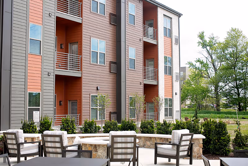 Outdoor seating area with cushioned chairs arranged around a stone fire pit in front of a multi-story residential building with balconies. Green shrubs and trees surround the area under a cloudy sky.