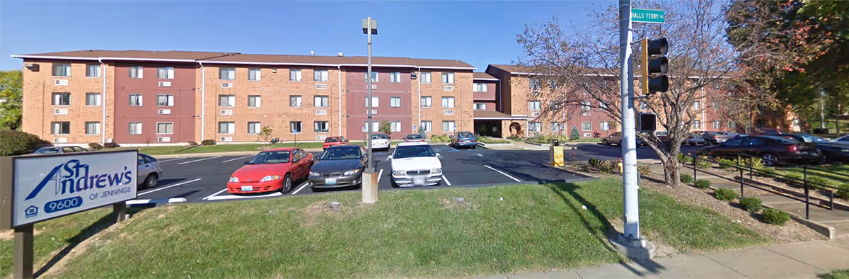 Exterior view of St Andrews of Jennings II senior living facility showing a multi-story brick building with several windows, a parking lot with cars, a streetlight, and a sign with the facility's name and address.
