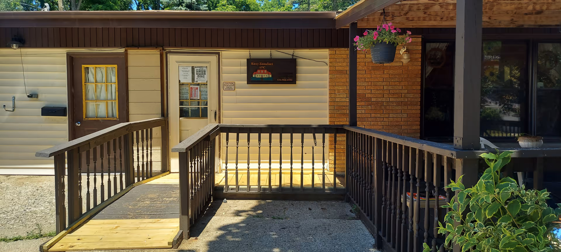 Entrance to Kozy Komfort Battle Creek AFC showing a wooden ramp with railings leading to a door. The building exterior has beige siding and brick walls. There is a hanging flower pot with pink flowers on the right side near the entrance.