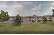 Exterior view of a single-story senior apartment building with a parking lot in front, several cars parked, and green grass with a few trees in the foreground under a partly cloudy sky.