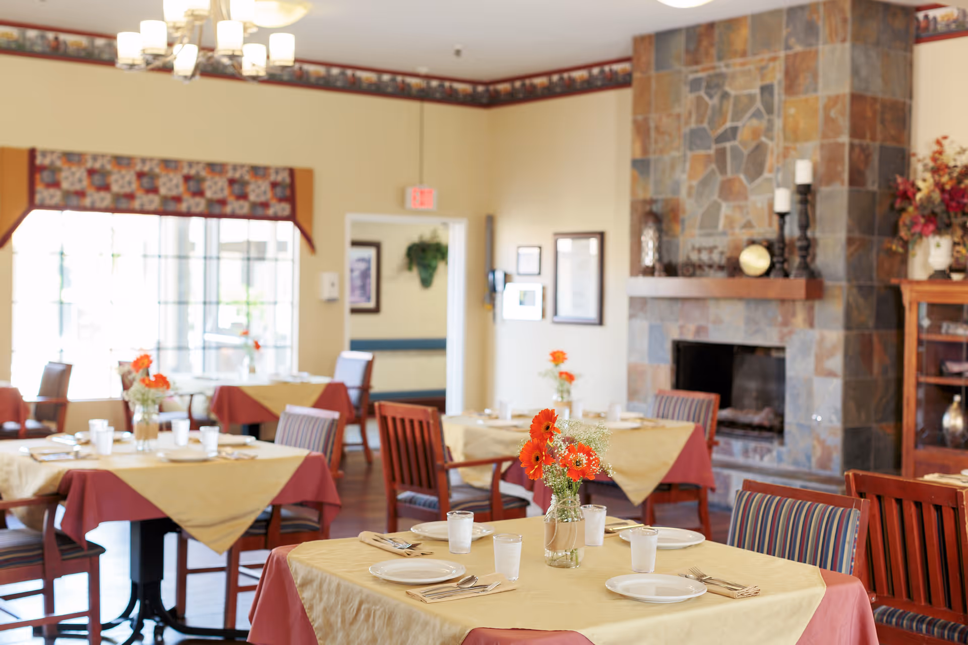 A cozy dining room with tables covered in beige and red tablecloths, each set with plates, glasses, and silverware. Vases with orange flowers decorate the tables. The room features a stone fireplace and large windows letting in natural light.