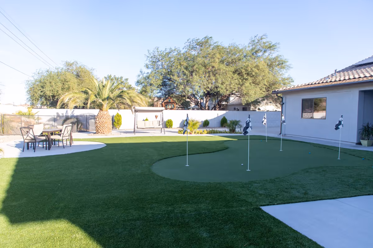 Outdoor area with a putting green featuring several golf holes with flags, a seating area with a table and chairs, a swing bench, palm trees, and other greenery, adjacent to a building with a tiled roof.