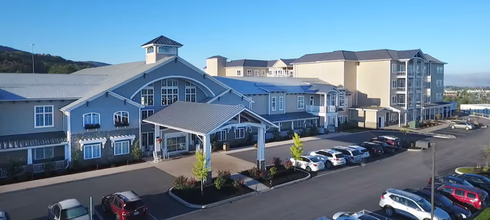 Exterior view of Highland Park Senior Living facility showing a large multi-story building with a covered entrance, multiple windows, and a parking lot with several cars parked. The building is surrounded by landscaped areas with small trees and shrubs, set against a backdrop of hills and clear blue sky.