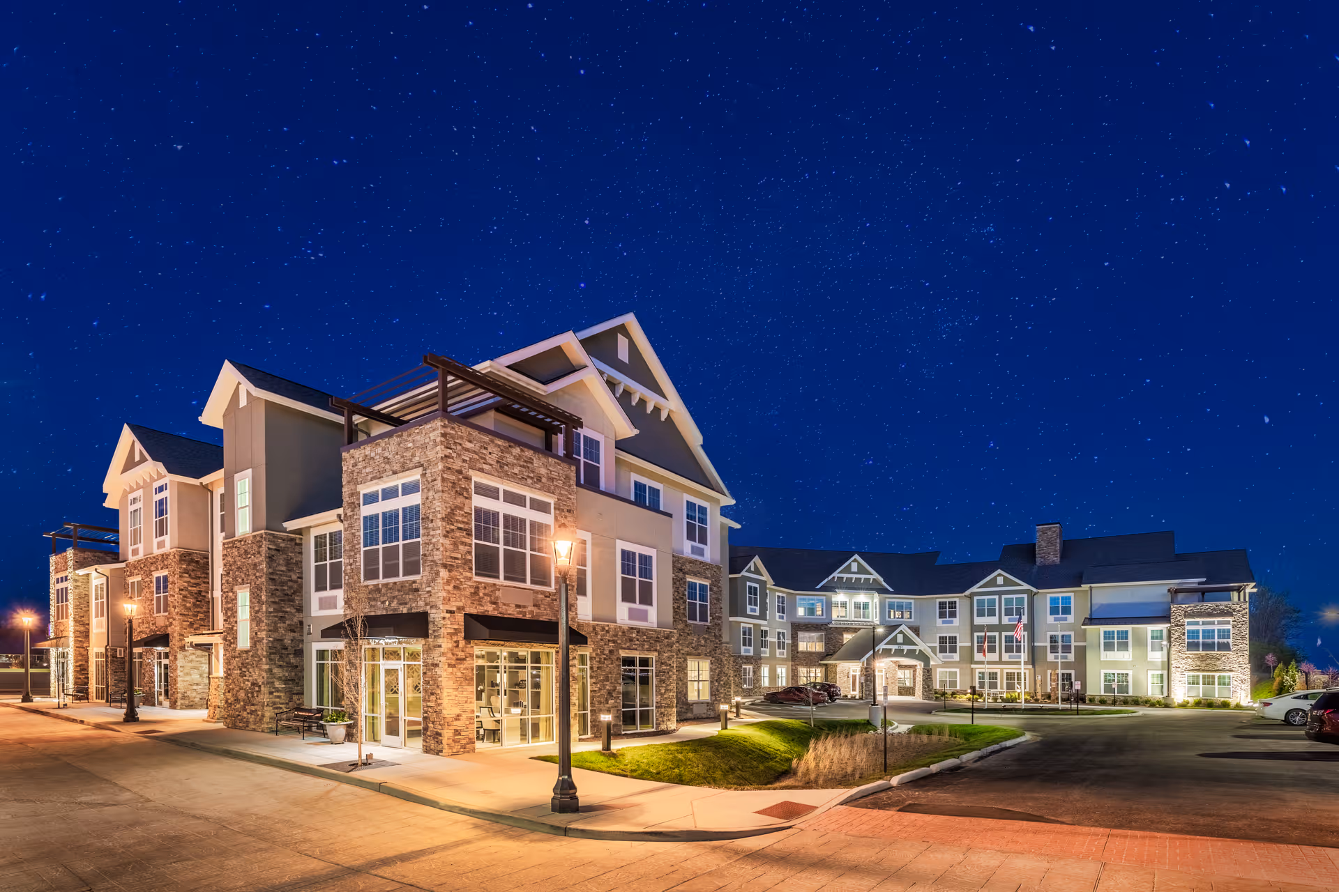 Nighttime exterior view of a large, well-lit senior living facility building with multiple floors, stone and siding facade, and a clear starry sky above.