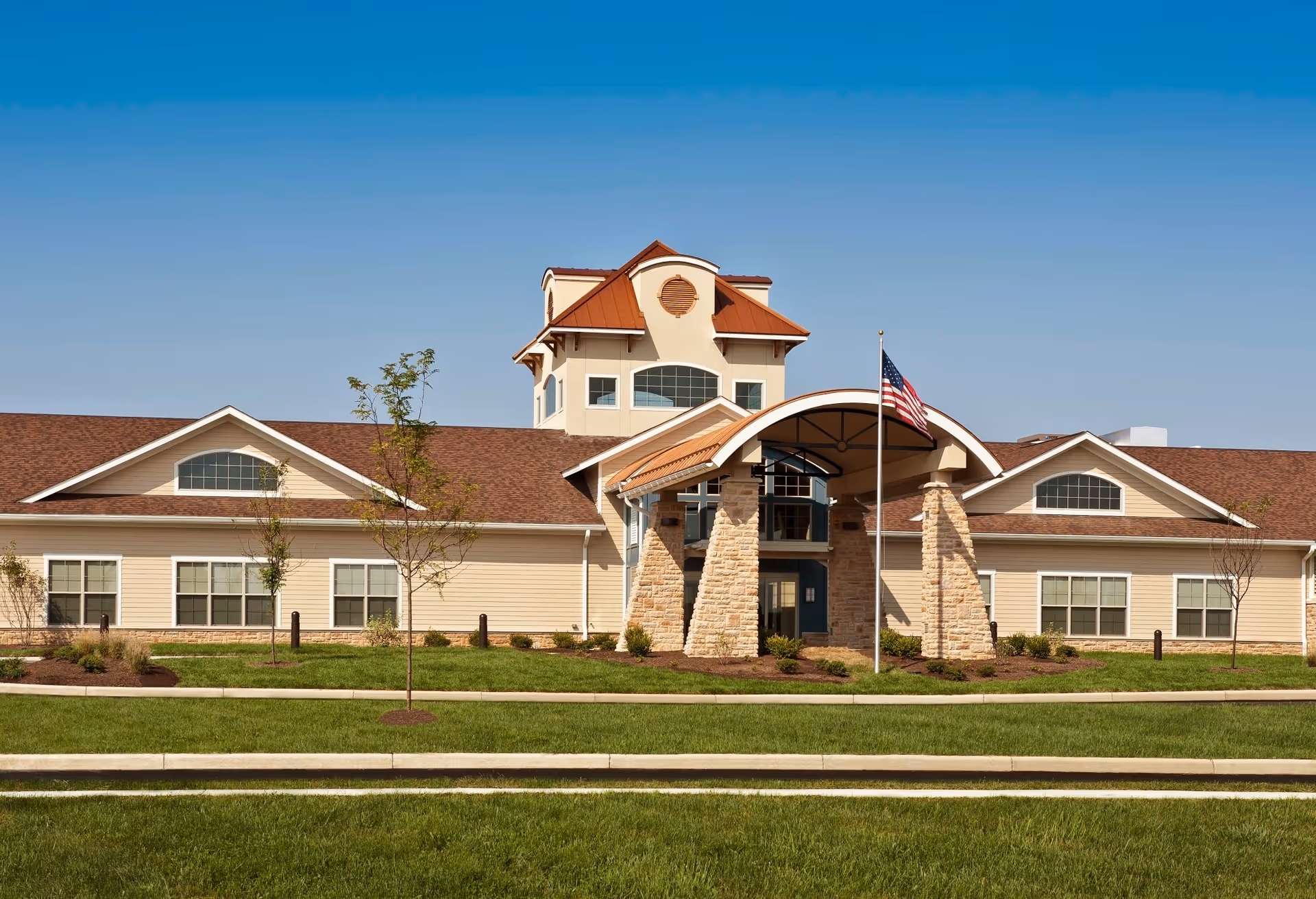 Front exterior view of a single-story building with a peaked roof and a central tower-like structure. The entrance features stone pillars supporting a curved roof, and an American flag is displayed on a flagpole in front. The building is surrounded by a well-maintained lawn and small trees under a clear blue sky.