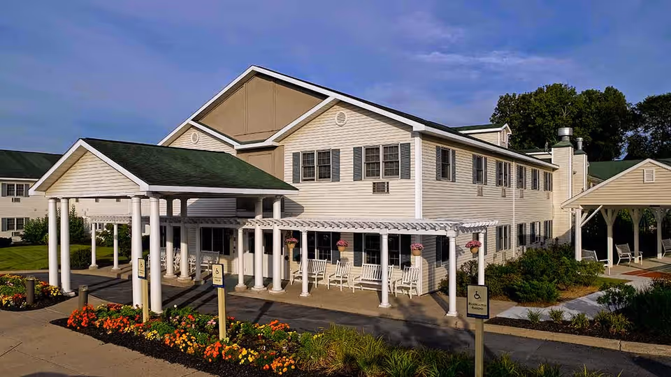 Exterior view of a two-story senior living facility building with beige siding and green roof. The entrance features a covered drop-off area supported by white columns, with white benches and hanging flower pots along the front porch. There are landscaped flower beds with colorful flowers and greenery around the entrance. The sky is clear with some clouds.