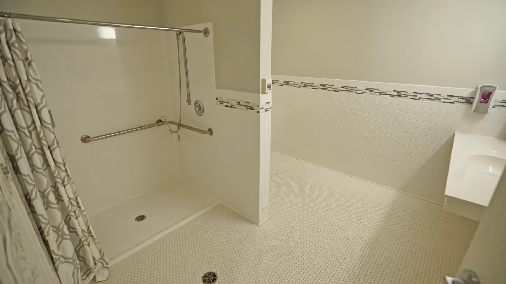 A spacious, accessible bathroom featuring a walk-in shower with grab bars and a patterned shower curtain. The walls are tiled halfway up with white tiles and a decorative horizontal strip. There is a white sink countertop visible on the right side.