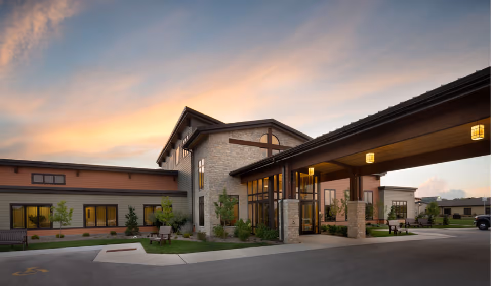 Exterior view of Pine Haven Christian Communities Haven Drive Campus building at sunset, featuring a covered entrance with stone pillars, large windows, landscaped greenery, benches, and a partly cloudy sky.