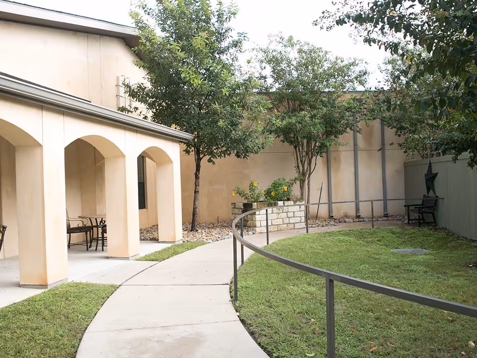 Outdoor courtyard area with a curved concrete pathway bordered by a metal railing, green grass, trees, and a beige building with arches and outdoor seating.