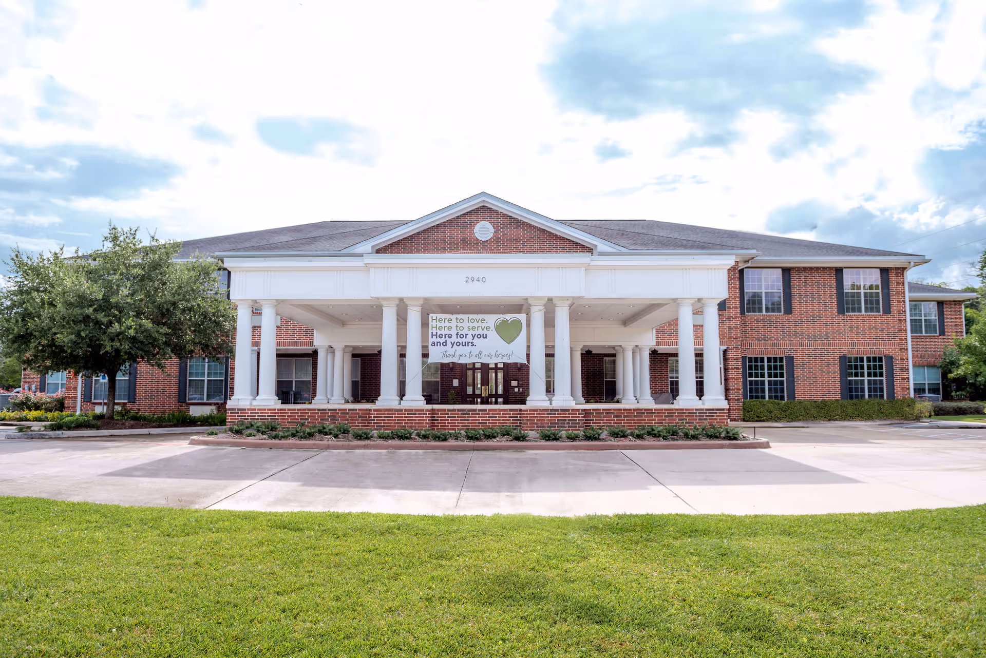 Front exterior view of The Reserve at Pearland Assisted Living and Memory Care building with a large covered entrance supported by white columns, brick facade, and a banner that reads 'Here to love. Here to serve. Here for you and yours.'