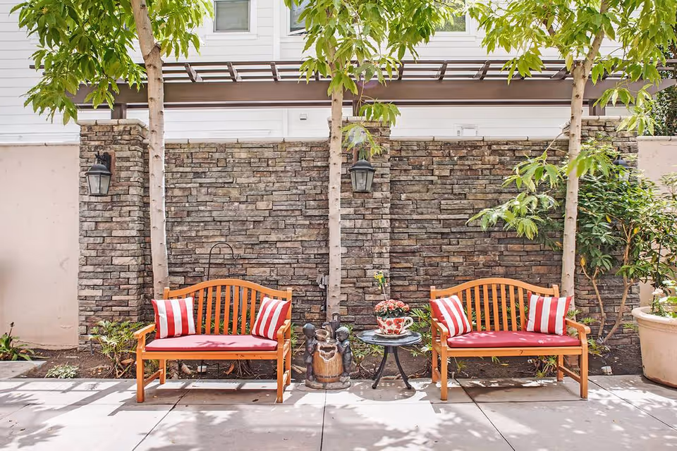 Outdoor seating area with two wooden benches featuring red cushions and red-and-white striped pillows, positioned on a paved patio in front of a stone wall with two trees and a small round table with a flower pot between the benches.
