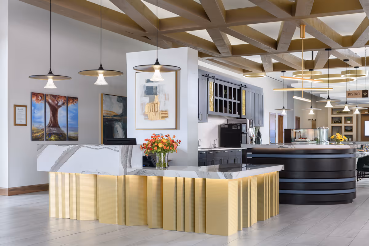 Modern reception area with a marble and gold-toned front desk, pendant lights hanging from a wooden beam ceiling, abstract artwork on the walls, and a bistro sign visible in the background.