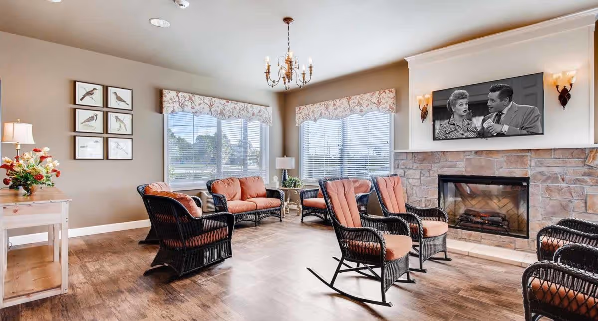 A cozy living room with wicker furniture including rocking chairs and sofas with orange cushions, a stone fireplace with a TV mounted above it showing a black and white scene, large windows with floral valances letting in natural light, wooden flooring, and wall decorations including framed bird pictures and a flower vase on a wooden table.