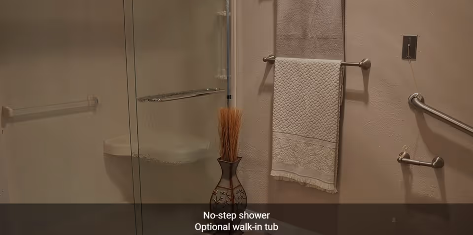 Bathroom with a no-step shower featuring a glass door, a towel rack with two towels, a decorative vase with dried plants, and grab bars on the wall for safety.