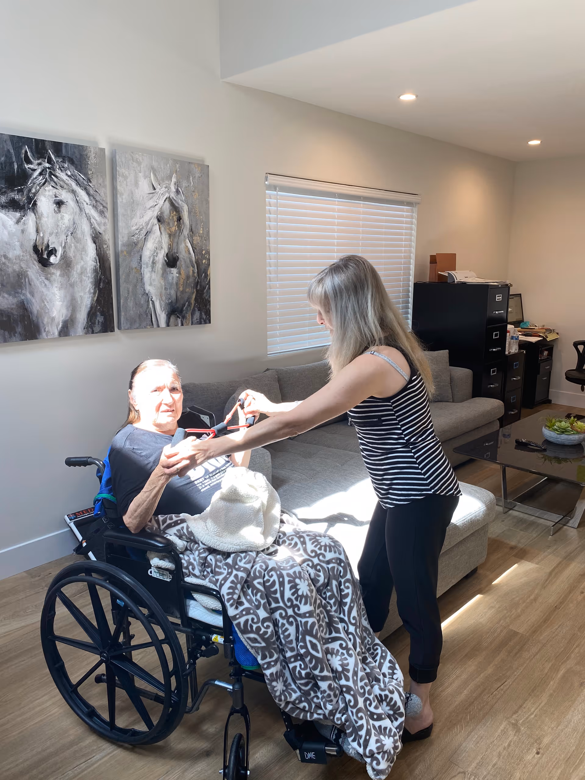 An elderly woman sitting in a wheelchair covered with a patterned blanket is being assisted by a younger woman in a striped tank top in a well-lit living room. The room has a gray sectional sofa, a glass coffee table with a plant, filing cabinets, and two paintings of white horses on the wall.