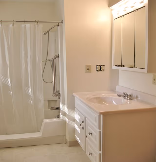 A clean and bright bathroom featuring a white shower with a curtain, a built-in grab bar, a white vanity with a sink, and a mirrored medicine cabinet above the sink. The walls and floor are light-colored, and there is a row of lights above the mirror.