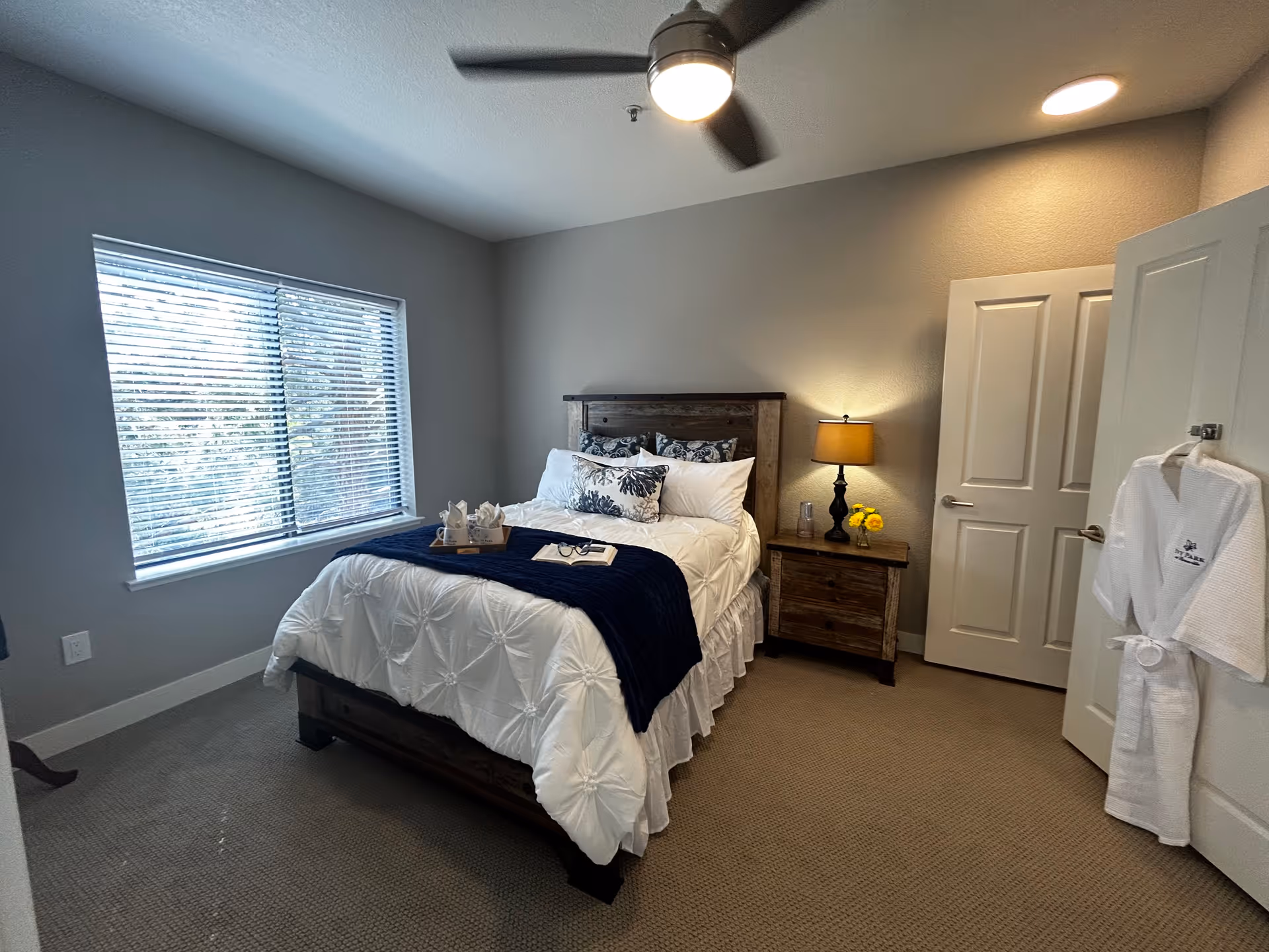 A cozy bedroom with a wooden bed frame and white bedding accented by a dark blue throw and decorative pillows. A wooden nightstand with a lamp and yellow flowers is beside the bed. A window with blinds lets in natural light. A white robe hangs on the open door, and a ceiling fan is visible above.