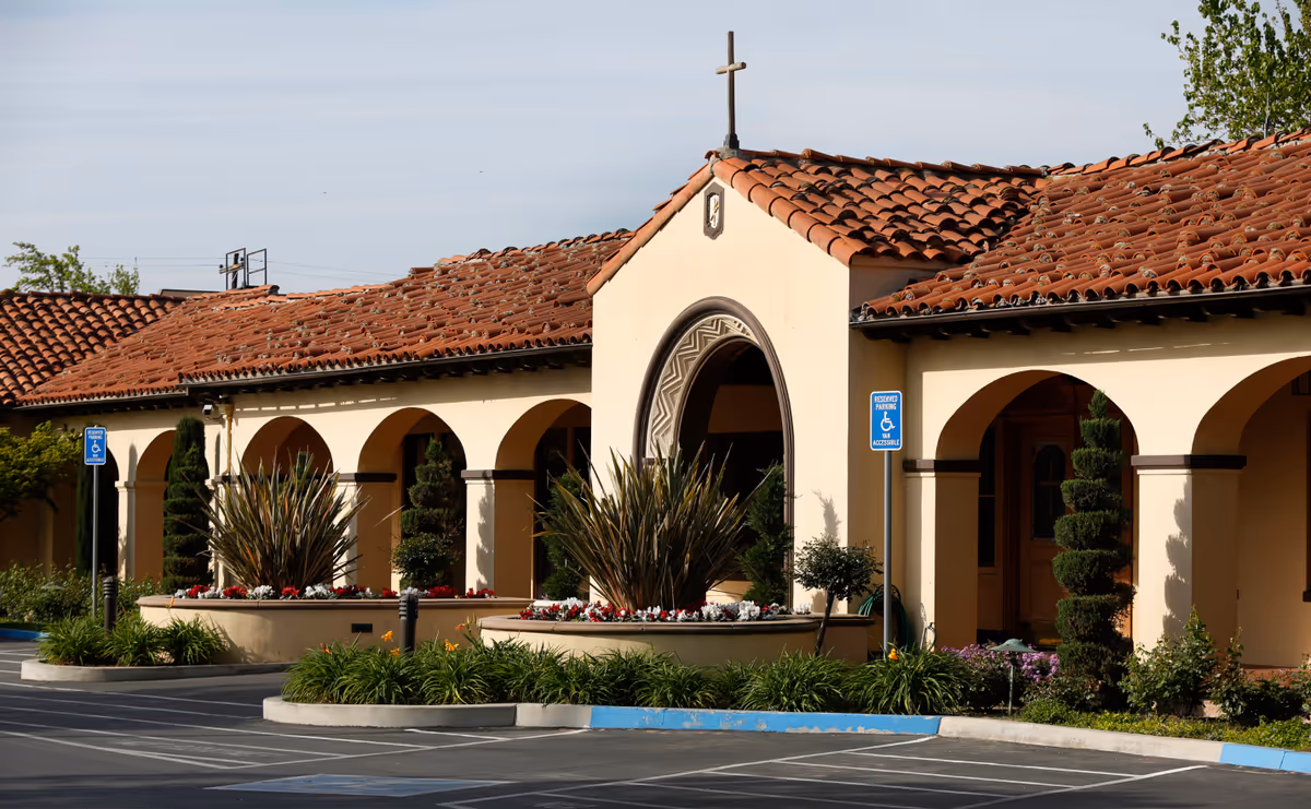 Exterior view of Nazareth House of Fresno featuring a building with a red tile roof, beige walls, arched doorways, and a cross on top. The foreground shows a parking area with reserved handicapped parking signs and landscaped plants and flowers.