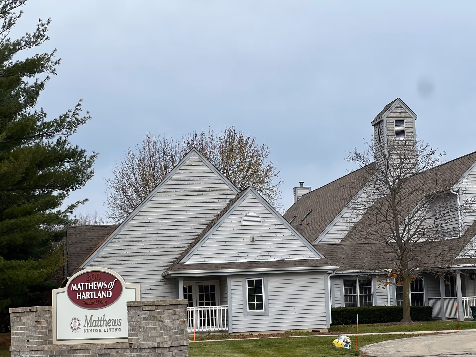 Exterior view of Matthews of Hartland senior living facility showing a light gray building with multiple peaked roofs, a small tower, and a sign in front that reads '300 Matthews of Hartland Senior Living' and 'Matthews Senior Living'. There are trees, some with no leaves, and a cloudy sky in the background.