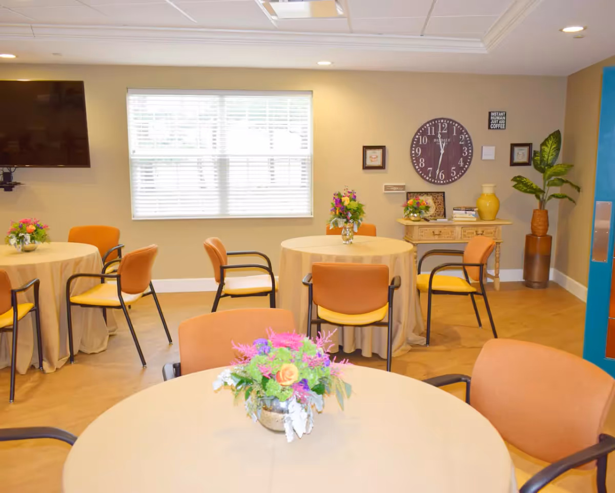 A bright and cozy dining room with round tables covered in beige tablecloths, each adorned with colorful floral centerpieces. Orange cushioned chairs surround the tables. A large window with white blinds lets in natural light. On the beige wall, there is a large round clock, small framed pictures, and a sign that reads 'INSTANT HUMAN JUST ADD COFFEE'. A wooden console table with decorative items and a tall potted plant are also visible. A flat-screen TV is mounted on the left wall.