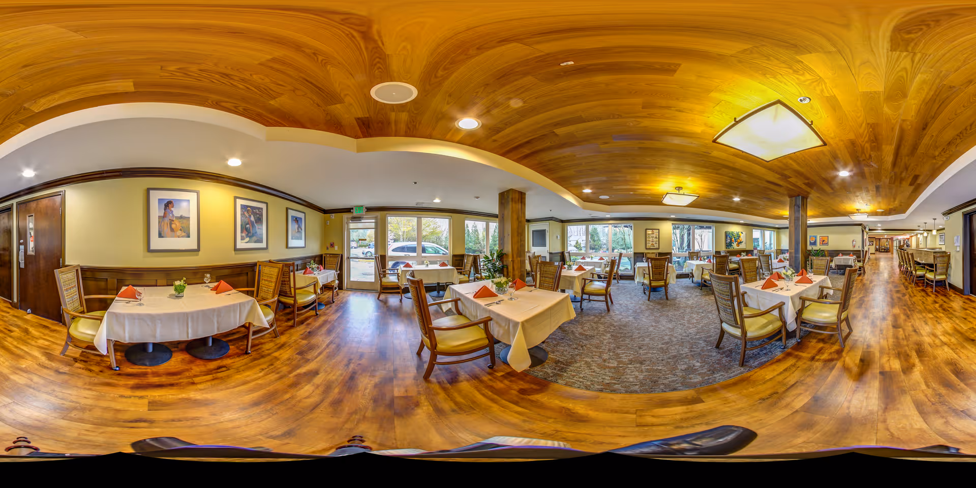 A spacious dining room in a senior living facility with multiple tables covered in white tablecloths and set with folded orange napkins. The room features wooden flooring, a wooden ceiling with recessed lighting, and large windows letting in natural light. There are framed artworks on the walls and comfortable wooden chairs with cushions around the tables.