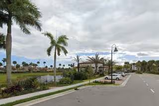 A paved road leading to a parking area with several parked cars, palm trees lining the street, a small pond with a fountain, and a building in the background under a cloudy sky.