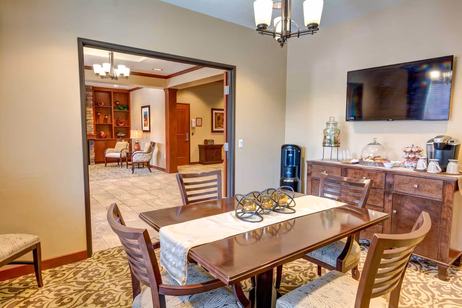 A cozy dining area with a wooden table and four chairs, decorated with a cream-colored table runner and three candle holders. On the right wall, there is a mounted flat-screen TV above a wooden sideboard that holds a water dispenser, a glass jar with fruit-infused water, a covered plate of pastries, and a coffee maker with cups. The room has beige walls and patterned carpet. Through an open doorway, a seating area with two upholstered chairs, a small table, and built-in wooden shelves with decorative items is visible.