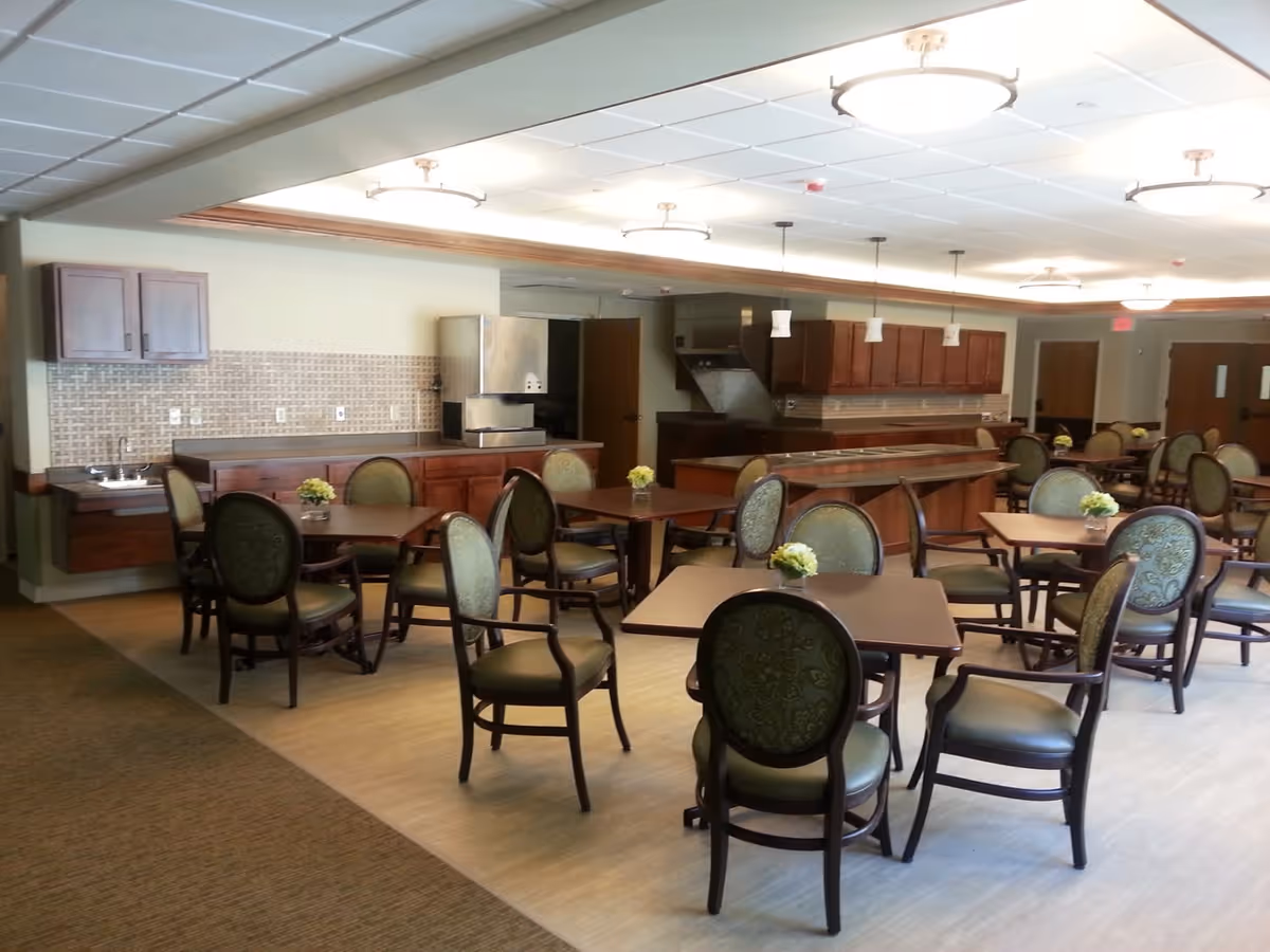 A spacious dining area with multiple square wooden tables and green upholstered chairs arranged neatly. The room features a kitchen area with wooden cabinets, a countertop, a small sink, and an ice machine. The ceiling has recessed lighting and hanging pendant lights above the kitchen counter. Small floral centerpieces are placed on each table.