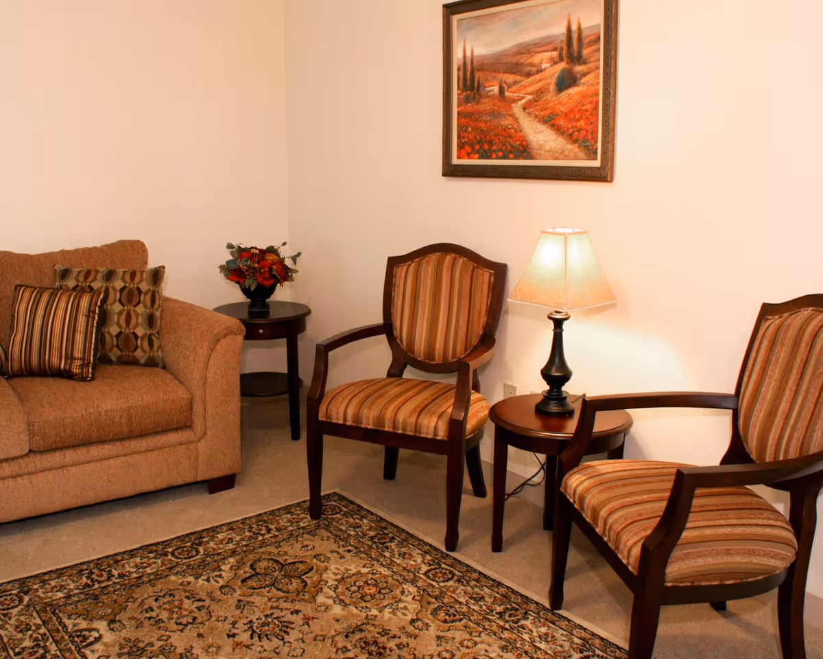 A cozy living room area with a brown upholstered sofa adorned with patterned cushions, two wooden armchairs with striped upholstery, a small round wooden side table with a lamp, another small table with a floral arrangement, and a framed landscape painting on the wall.