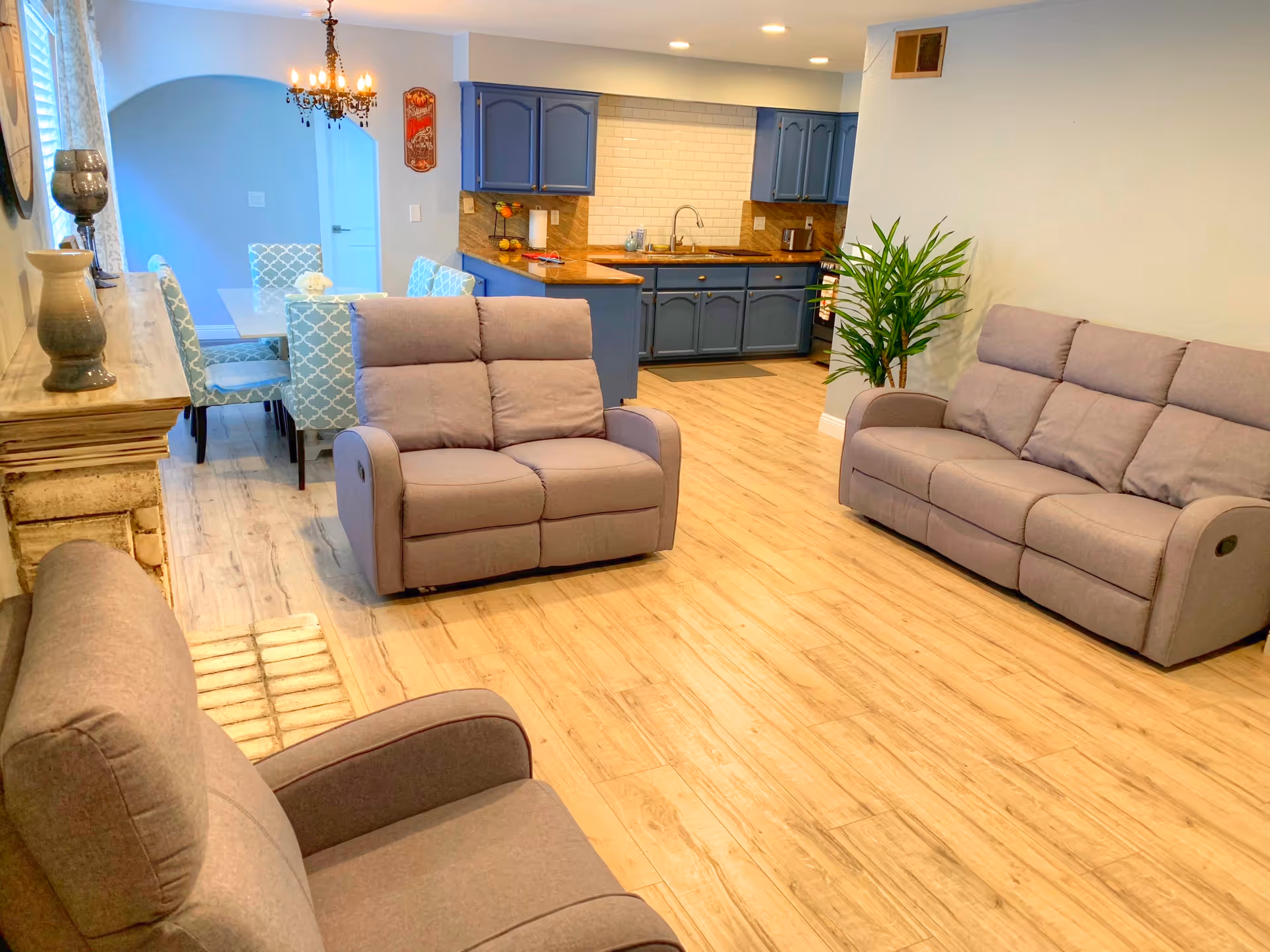 A bright and spacious living area featuring three gray reclining sofas arranged around a wooden floor. In the background, there is a dining area with a glass table and patterned blue chairs, and a kitchen with blue cabinets, a wooden countertop, and a white tiled backsplash. A green potted plant is placed near the wall, and a chandelier hangs above the dining table.
