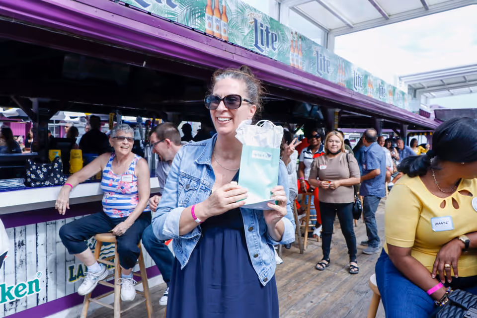 A group of people socializing at an outdoor bar area. A woman in the foreground wearing sunglasses and a denim jacket is smiling and holding a gift bag. Other people are sitting and standing around the bar, which has advertisements for Miller Lite and Heineken. The setting appears lively and casual with a wooden floor and a partially covered roof.