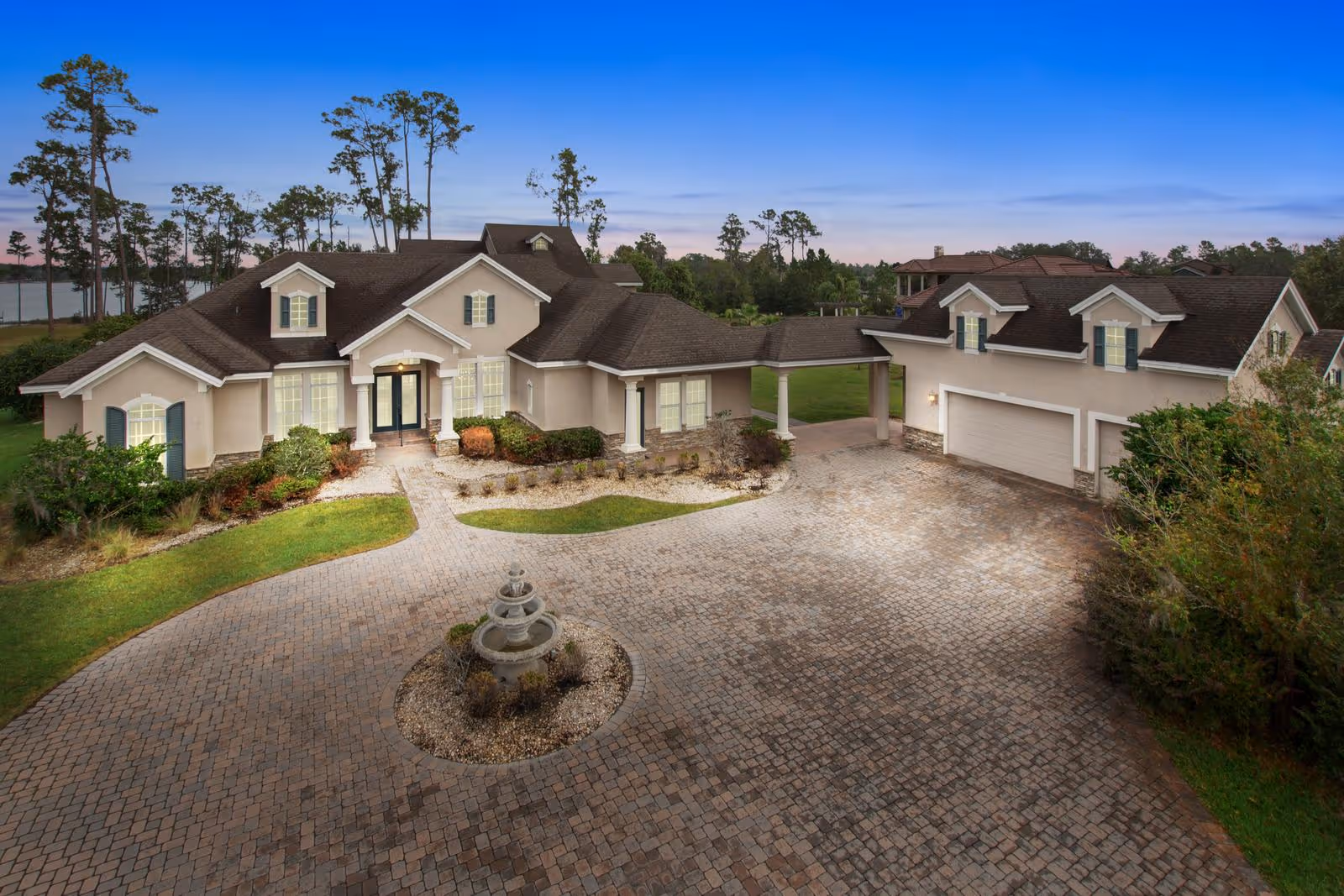 Exterior view of a large, elegant assisted living facility named Lake Hendon Estate Assisted Living, featuring a spacious driveway with a central fountain, well-maintained landscaping, and a backdrop of trees and a lake under a clear evening sky.