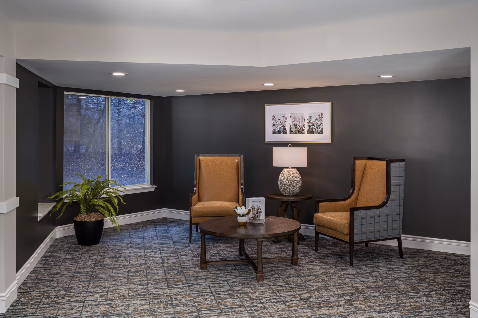 A cozy seating area in a senior living facility with two high-back armchairs upholstered in tan and plaid fabric, a round wooden coffee table with a small plant and a brochure on it, a side table with a decorative lamp, a framed floral artwork on a dark gray wall, a large window showing a wooded outdoor scene, and a potted fern plant on the carpeted floor.