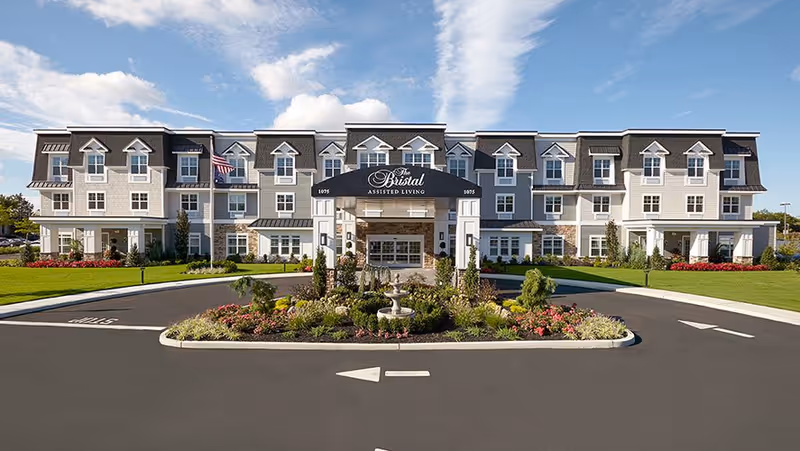 Front exterior view of The Bristal Assisted Living at Bethpage, a large three-story building with multiple windows and a covered entrance. The building is surrounded by well-maintained landscaping with green lawns, shrubs, and flowers. An American flag is visible near the entrance under a partly cloudy blue sky.